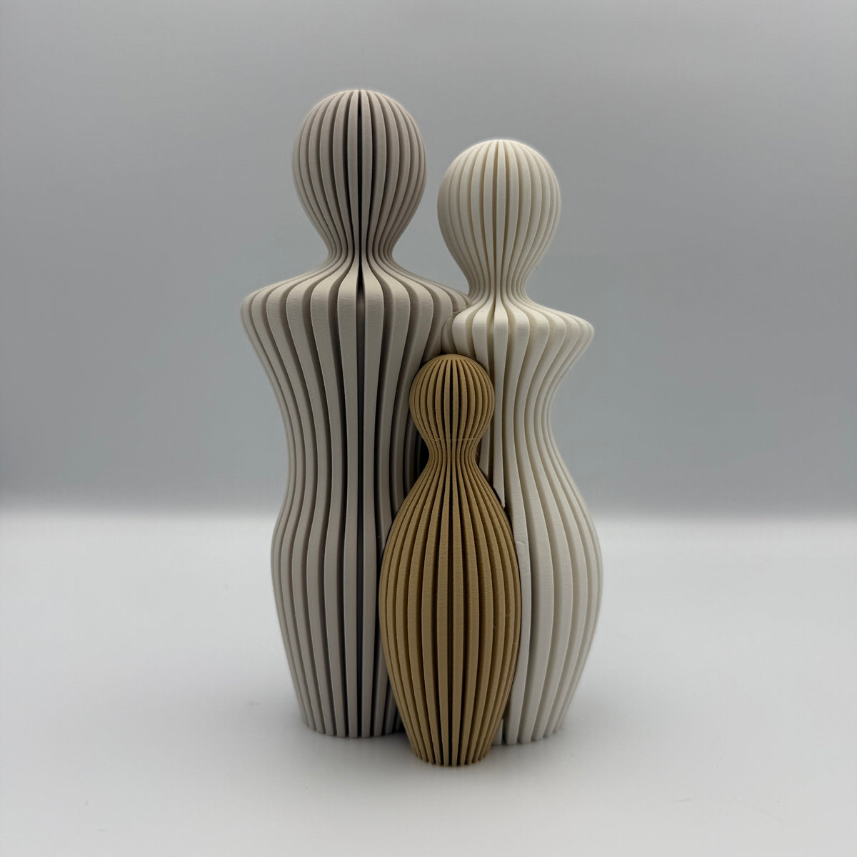 Familien, 3D-printet figur med to lyse voksne og ét gyldenbrunt barn i ribbet design.
